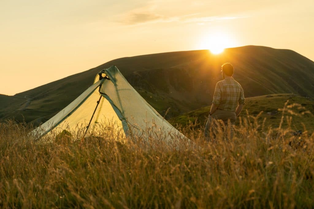 Man camping in mountains watches sunset beside tent