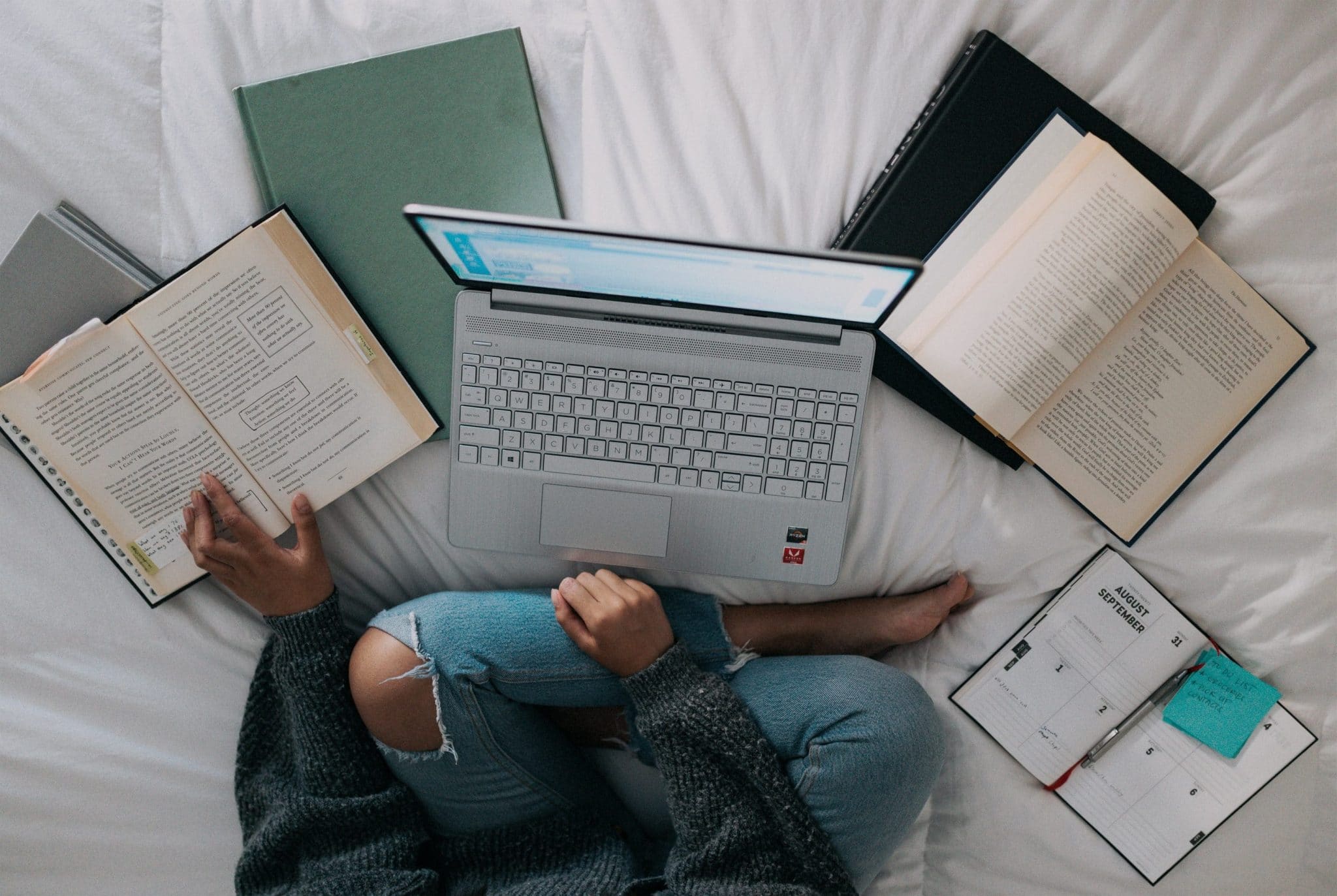 Woman sitting on bed surrounded by books for research, her laptop, and a planner.