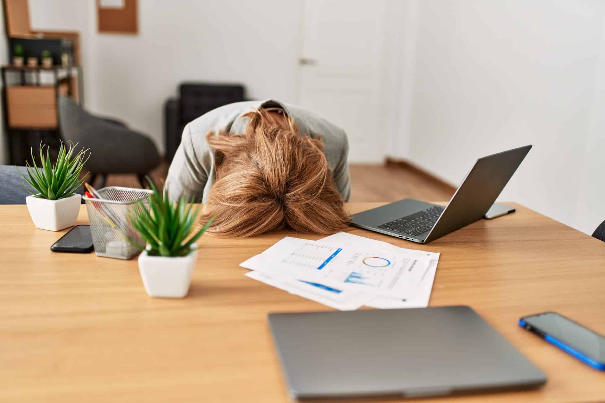 A woman rests her head on her work table. This scene underscores the importance of managing workloads and prioritizing well-being to prevent burnout.