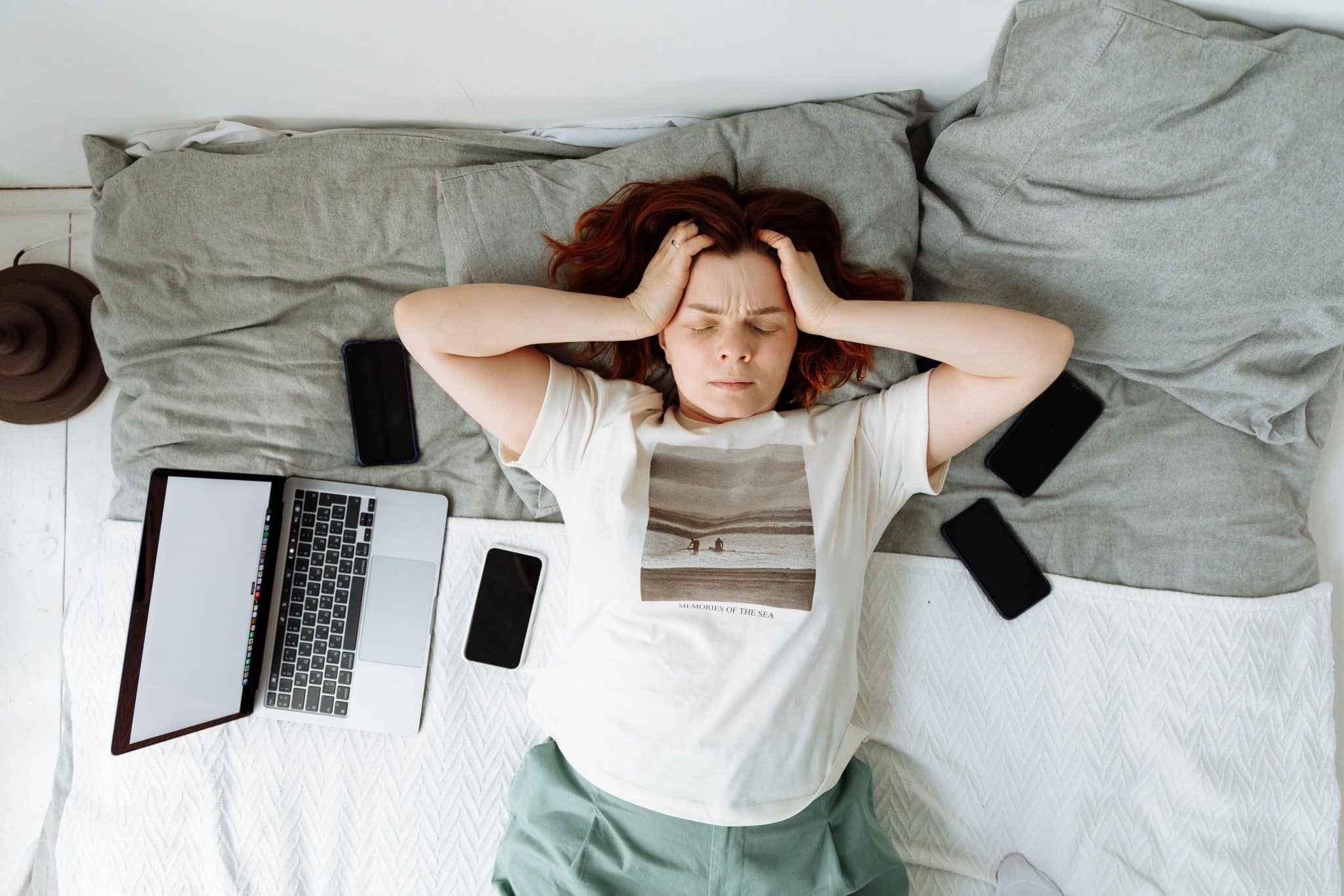 Stressed woman lying on bed surrounded by phone and laptop, illustrating cortisol spikes caused by screen overload.