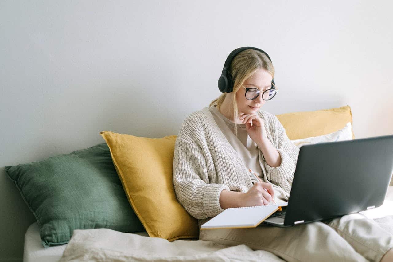 Woman practicing soft discipline while relaxing on a bed using a laptop and taking notes in a calm and cozy home workspace